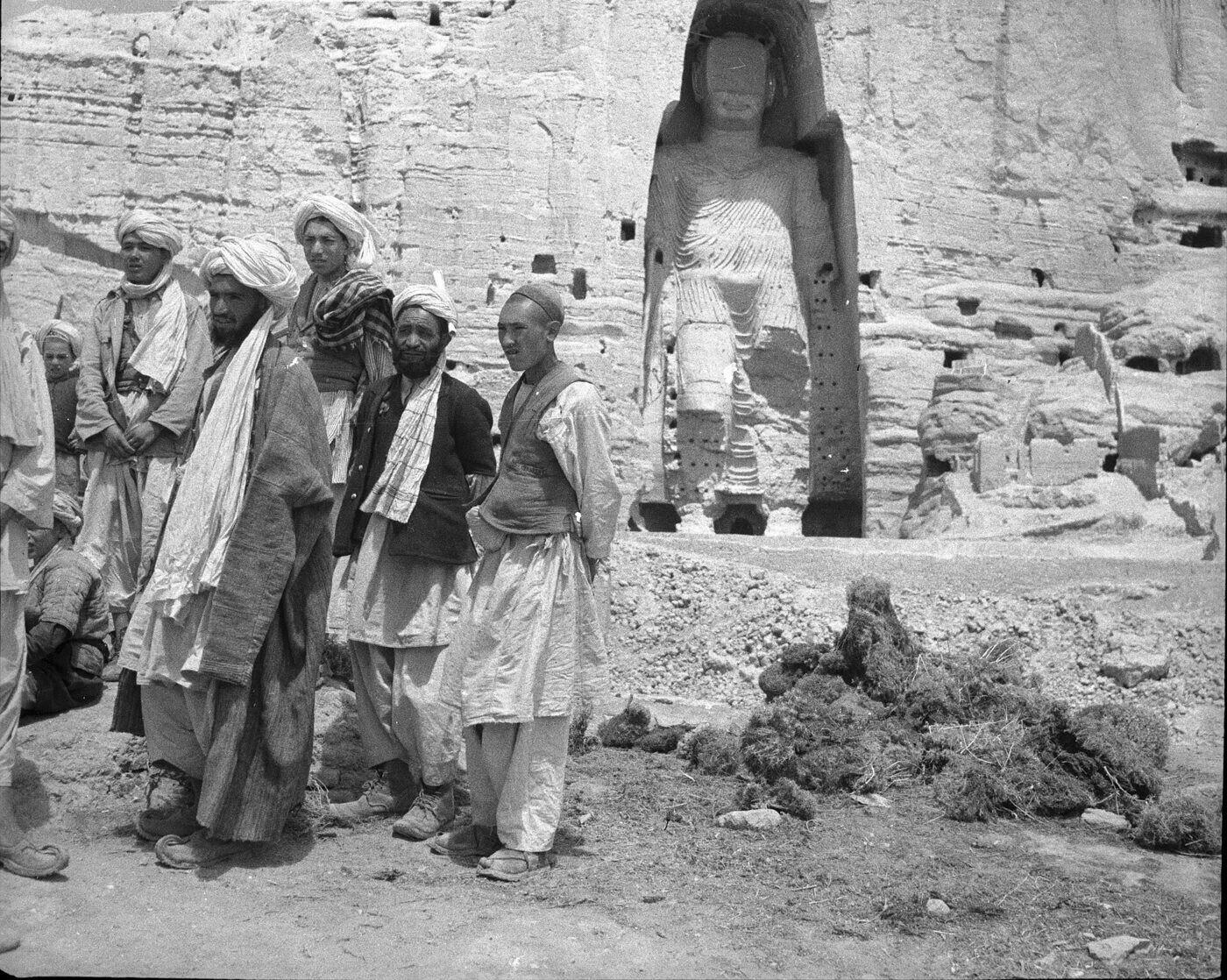 Black-and-white photograph of a group of men in turbans, caps and long coats standing on rough ground before a sandstone cliff, with a large standing Buddha figure carved into a niche in the cliff behind them and rock-cut caves around it.