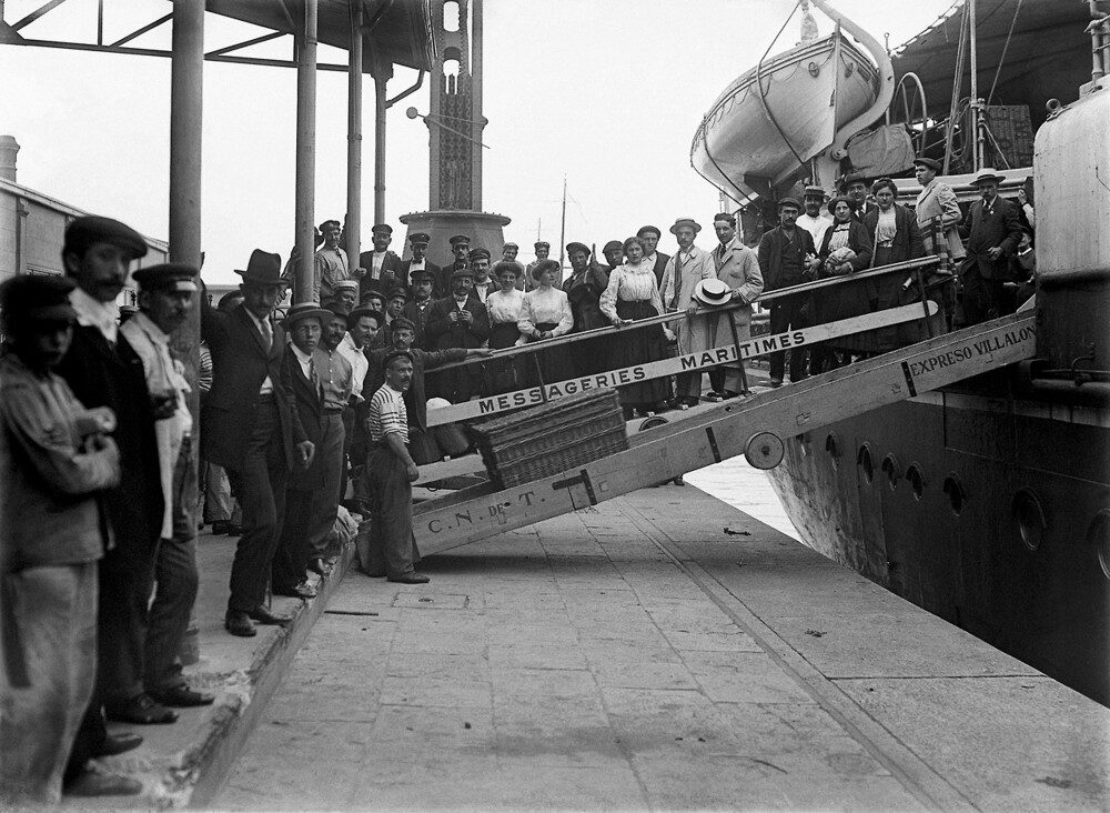 Archival photograph of passengers crowded on the gangway of a steamship, with men waiting on the quayside below.