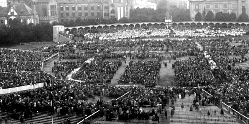 An elevated black-and-white photograph of a very large open-air gathering: massed singers fill an esplanade and the tiered stands around it, facing a long low-roofed wooden stage running the width of the ground, with trees and city buildings behind.