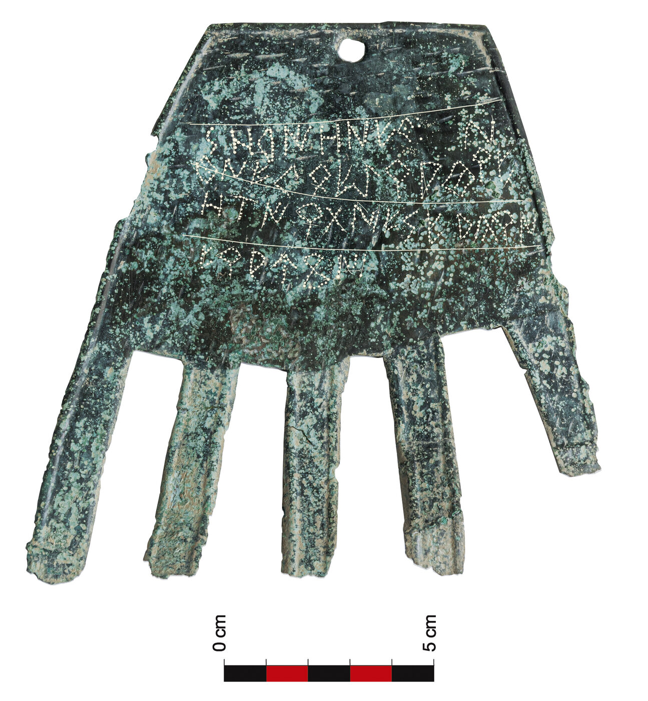 A corroded green bronze plate cut in the outline of a right hand with the fingers extended, photographed against white; four lines of angular dotted letters run across the palm, and a black-and-red scale bar marked nought to five centimetres lies beneath it.