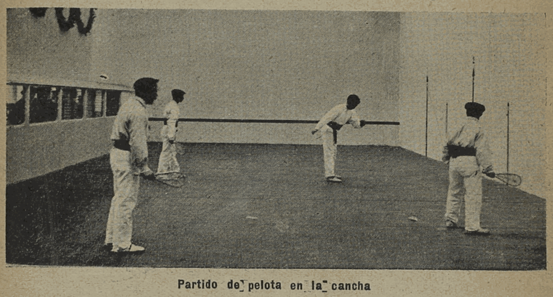 A 1904 halftone photograph of a pelota match: four players in white shirts and trousers, most of them holding stringed rackets, on either side of a cord strung across a walled court, with a railed spectator gallery along the left-hand side.
