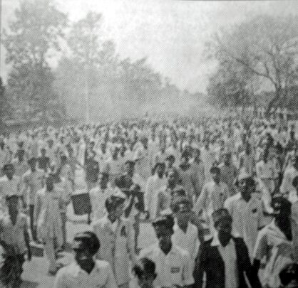 Black and white photograph of a large crowd of men in white clothing filling a tree-lined road, walking towards the camera.