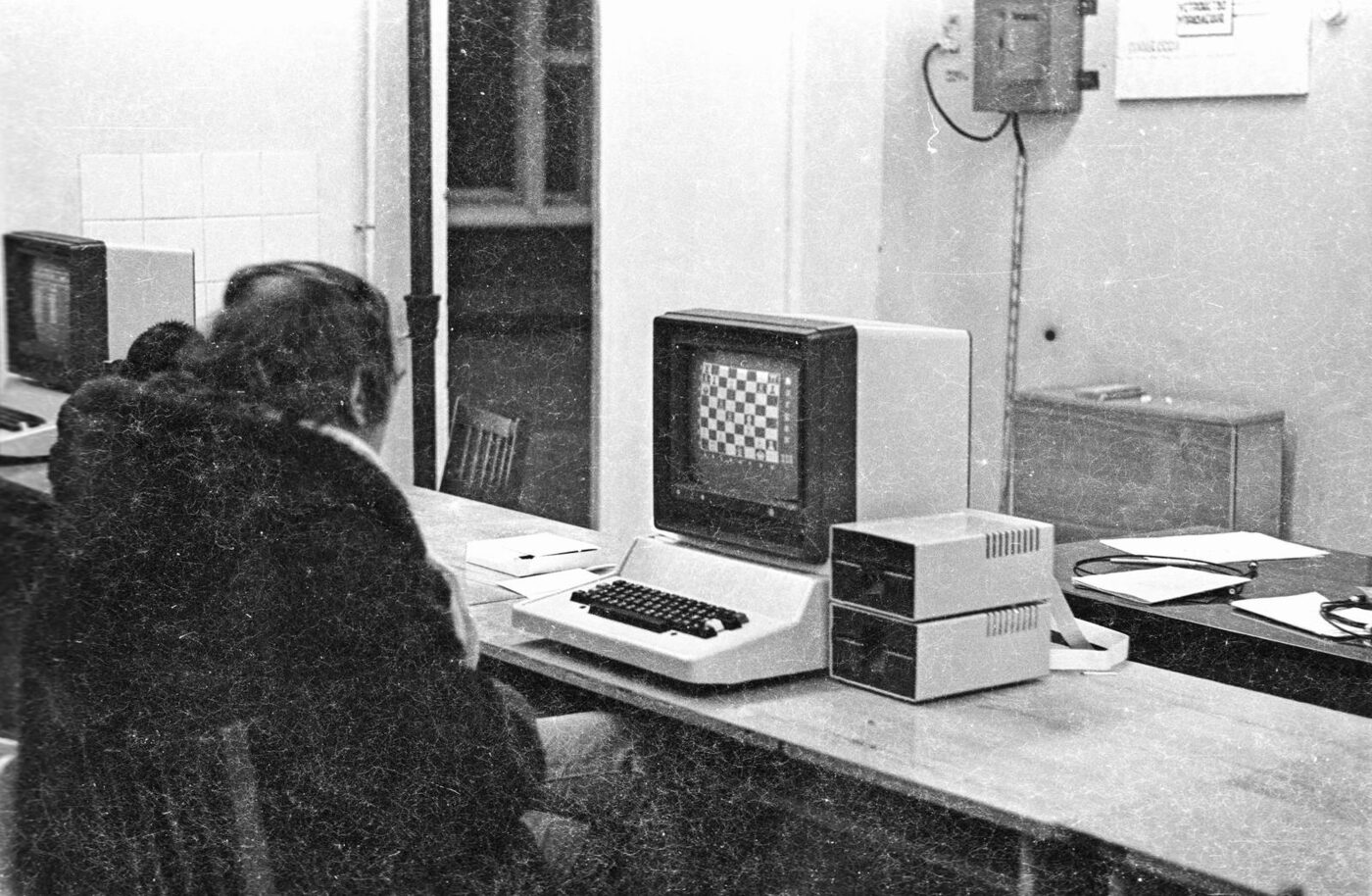 A grainy black-and-white photograph of a classroom: a wedge-shaped microcomputer with a separate monitor showing a chessboard and two disk drives stacked beside it; two people sit with their backs to the camera at another machine on the left.