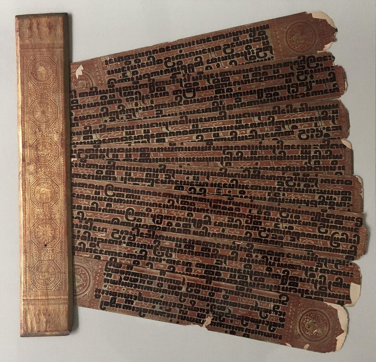 Several long narrow lacquered manuscript leaves fanned out, each bearing heavy square black letters on a red and gold ground, beside a gilded decorated binding board.