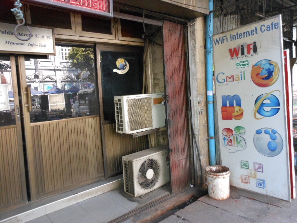 A street-level shopfront with a large signboard reading WiFi Internet Cafe beside browser and webmail logos, a smaller Myanmar Info-Tech public access centre sign to the left, and two air-conditioning units bolted beside the doorway.