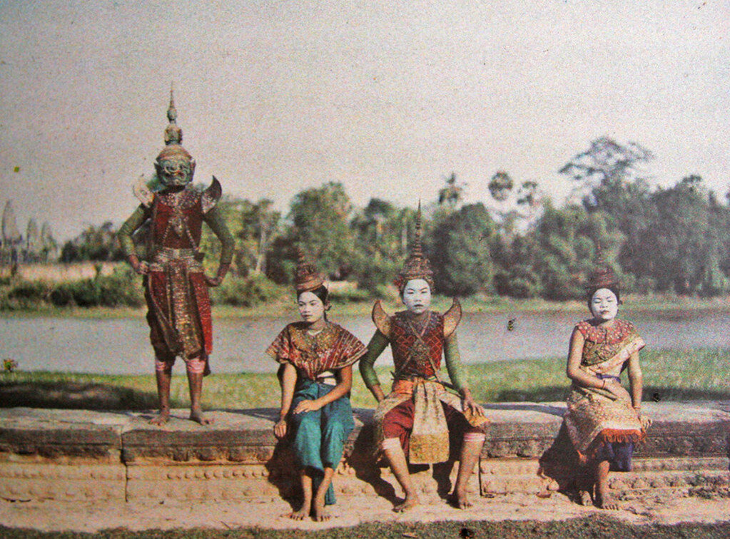 Early colour photograph of four Cambodian classical dancers in silk costume and pointed headdresses, three seated on a low stone terrace by water and one standing in a masked giant's costume.