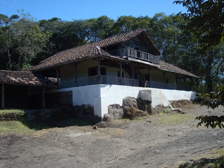 A two-storey colonial hacienda house with a tiled roof, timber verandah and whitewashed stone base, standing on bare ground at the edge of woodland under a clear sky.