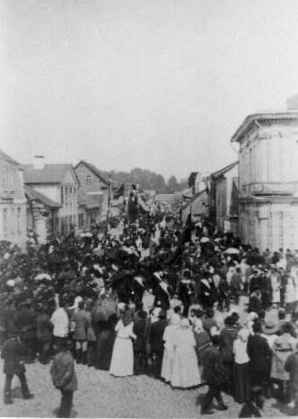 Black and white photograph of a crowd filling a wide unpaved street between low houses, with a decorated procession moving through the middle of it.