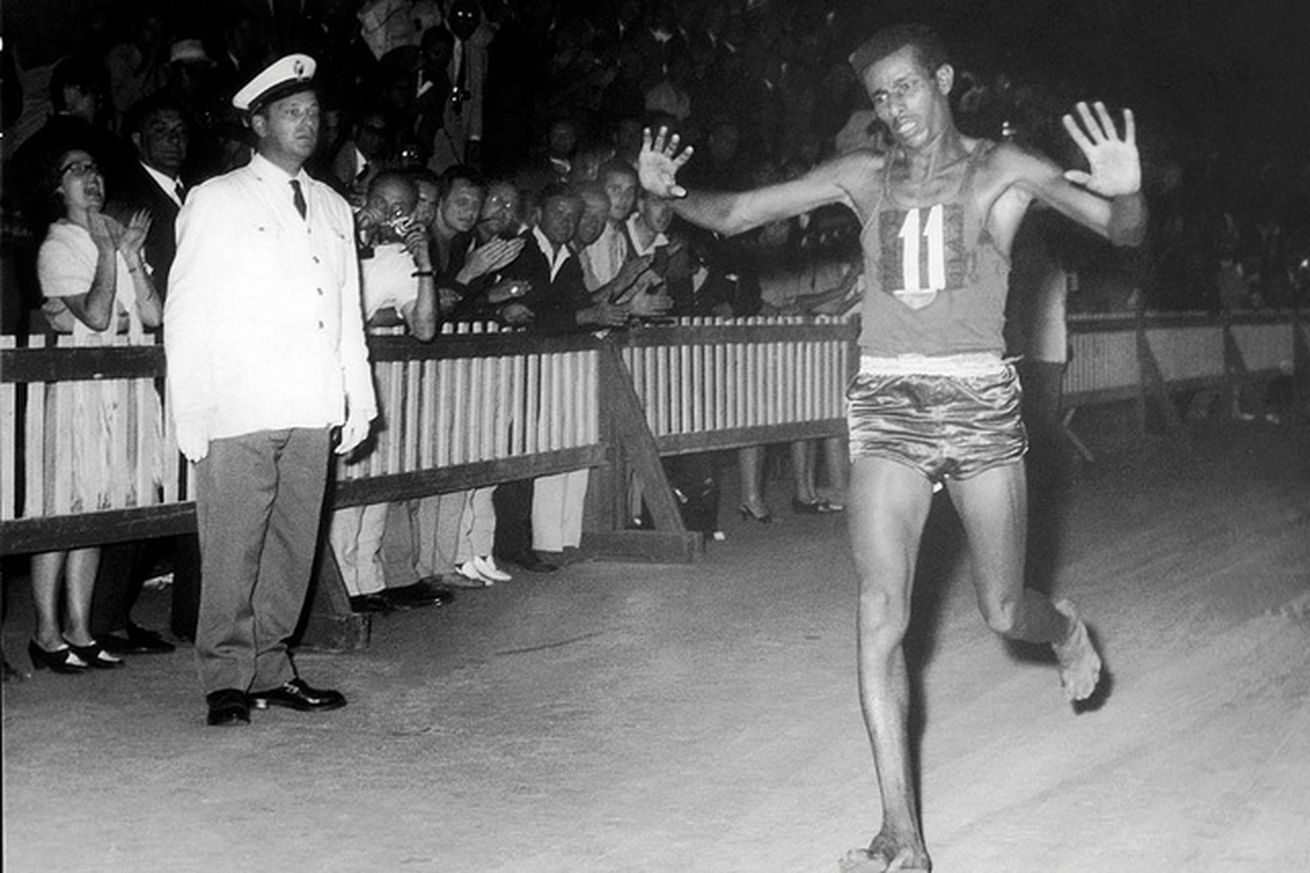 A black and white photograph of a lone barefoot runner in a dark singlet on a floodlit road, with spectators and officials lining the route behind him.