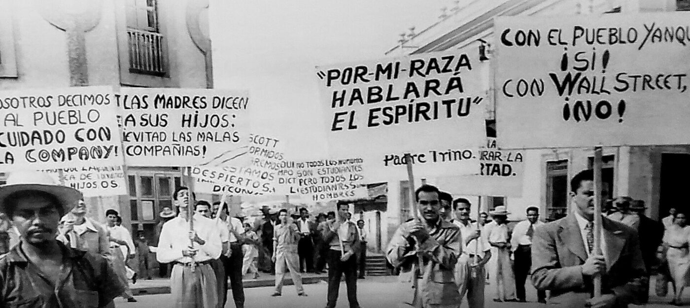 Black and white photograph of a street demonstration: men in shirtsleeves and hats carry large hand-lettered placards in Spanish above a crowd, with a two-storey building behind them.
