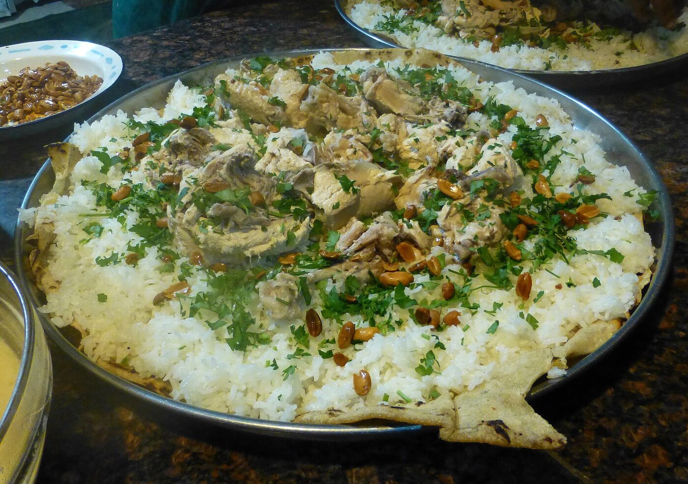 A large round metal tray of white rice topped with pieces of cooked meat, chopped parsley and fried nuts, resting on flatbread, with a second tray and small side dishes nearby.