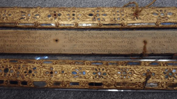 Close view of a palm-leaf manuscript from Laos, one inscribed leaf visible between wooden covers decorated with lacquer, gilding and inlaid mirror glass.