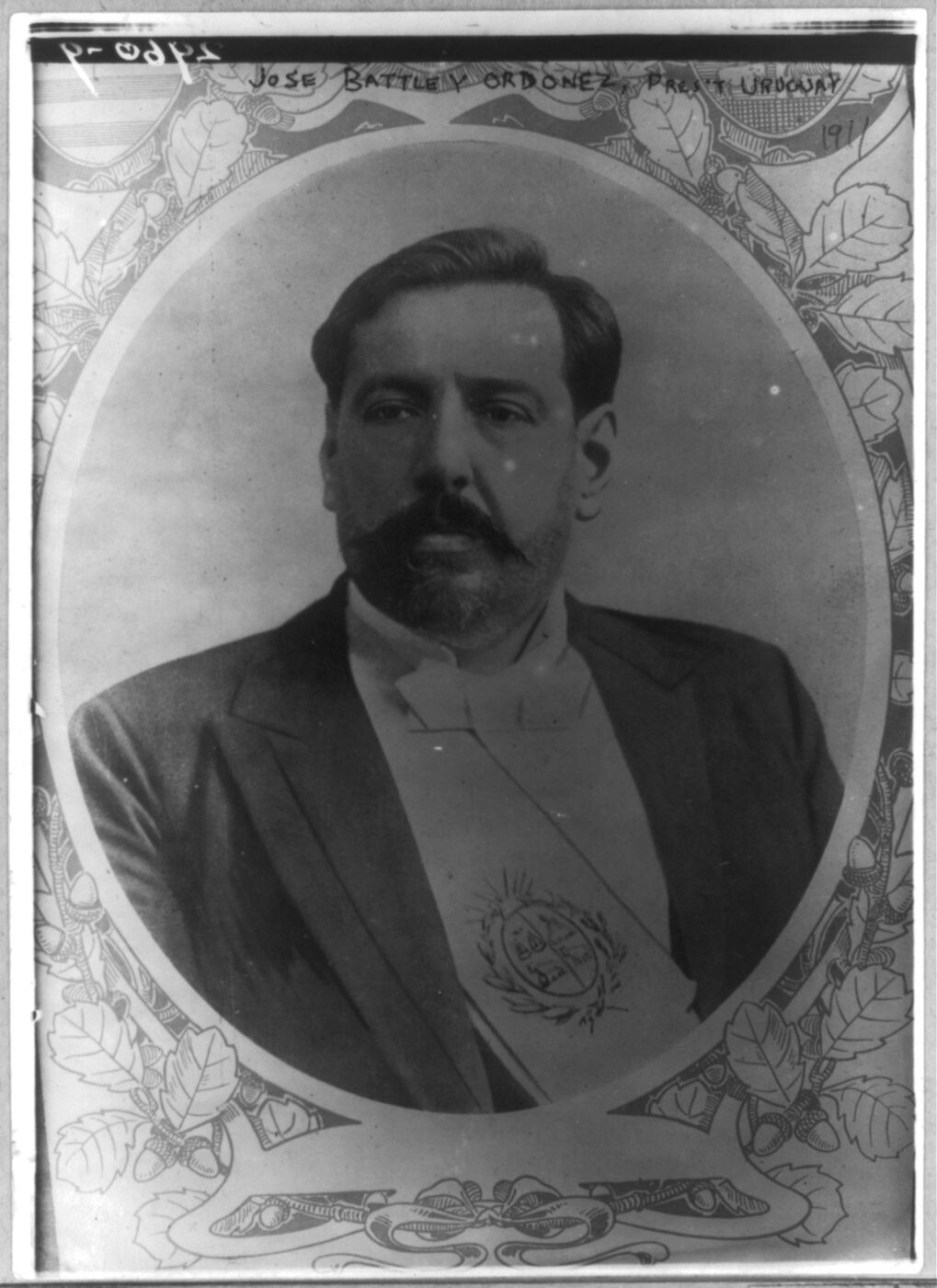 Black and white photograph of a moustached man in a dark suit wearing a presidential sash bearing the Uruguayan coat of arms, shown in an oval vignette within an ornamental printed border.