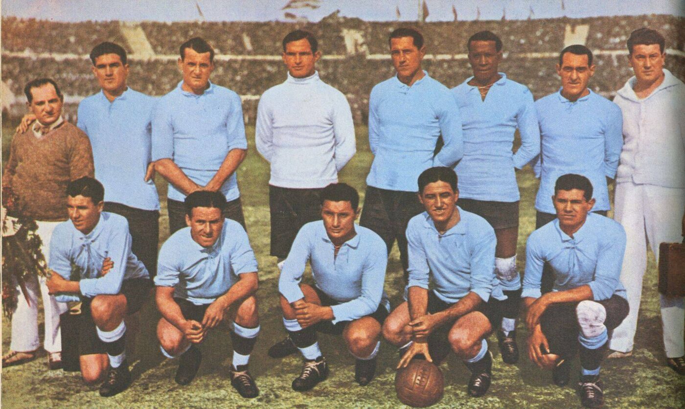Colour-tinted team photograph on a pitch before a packed stand: a back row in pale blue and white jerseys beside a man in a brown cardigan, and a front row crouching with a leather ball.