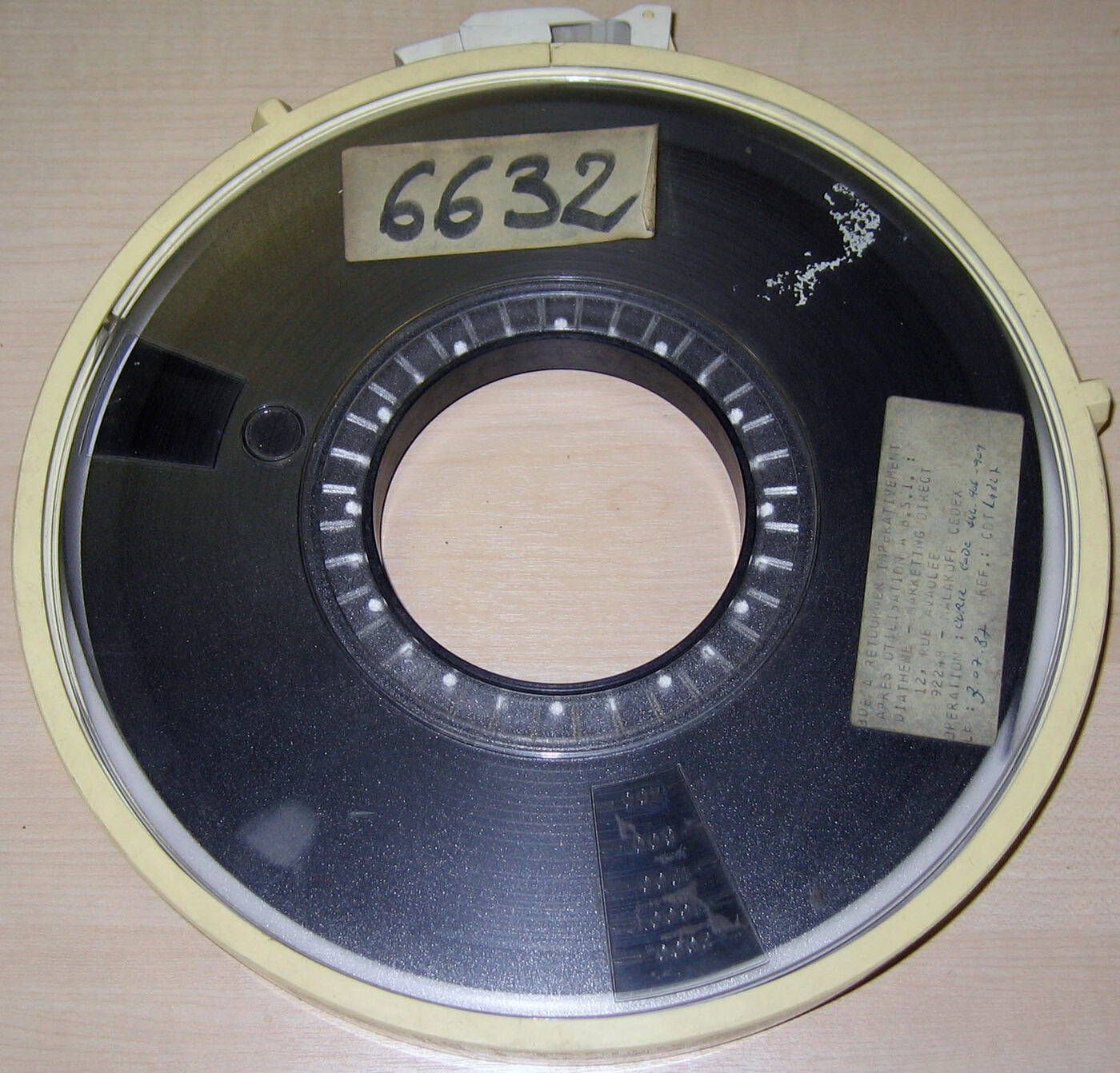 A reel of dark magnetic computer tape inside a circular plastic protective ring resting on a pale desk, with a stuck-on label reading 6632 near the top and a smaller handwritten label to the lower right.