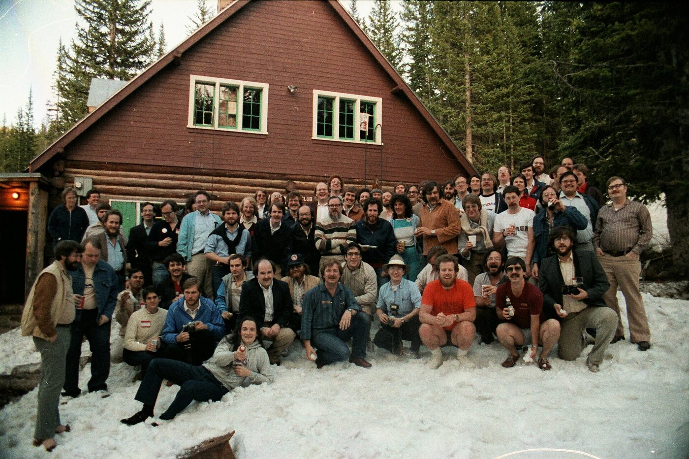 Colour photograph of some eighty people, mostly men in early-1980s casual dress, posed in rows on snow-covered ground in front of a dark red log building with a steep gable, conifers behind.