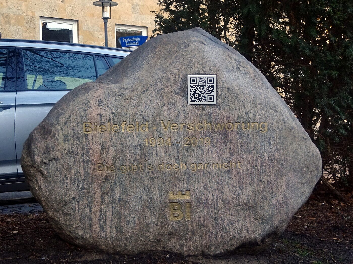 A large granite boulder standing on bare ground beside a parked car and a stone building, engraved in gold lettering with the words Bielefeld-Verschwörung, the dates 1994 to 2019 and a short German sentence, with a small square QR-code plaque set into the stone above the lettering and the city’s BI emblem below it.