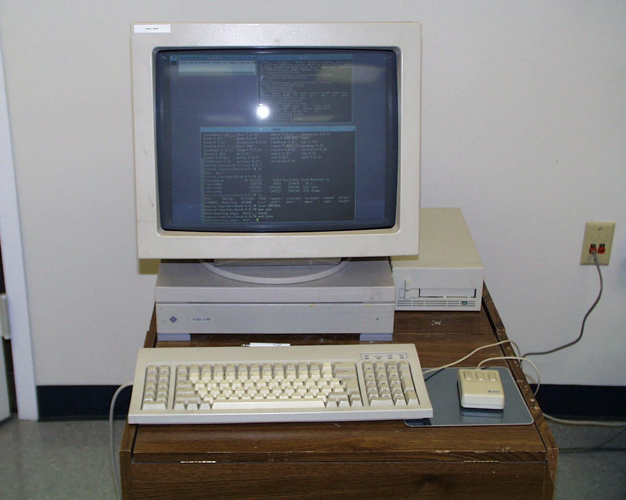 A late-1980s Unix workstation on a wooden desk: a large beige CRT monitor showing pale terminal windows of text, standing on a low beige system unit labelled Sun 3/80, with a beige keyboard and a three-button mouse on a pad in front and a small cartridge tape drive to the right.